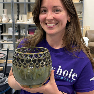 A member holding a round green vessel with a cut-out scale pattern around the top and drippy glaze below at The Clay Hole pottery studio in Draper, Utah