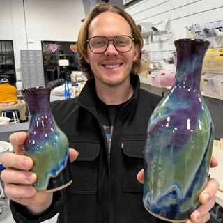 A member holding two vases, a small one and a tall one, with glossy purple, green and blue drippy glaze at The Clay Hole pottery studio in Draper, Utah