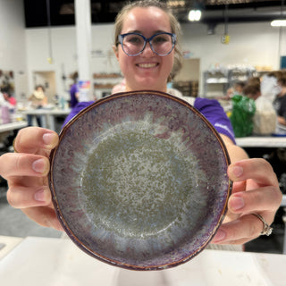 A member holding up a shallow round bowl with purple edges and a speckled gray-green center at The Clay Hole pottery studio in Draper, Utah