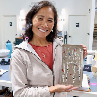 A member holding a rectangular taupe dish with white birch trunks and small red and blue birds at The Clay Hole pottery studio in Draper, Utah