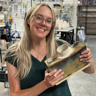 A member holding a rectangular vessel with glossy brown, gold and blue glaze and a round speckled opening on top at The Clay Hole pottery studio in Draper, Utah