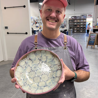 A member in an apron holding a round bowl with a pale cell pattern inside and a pink rim at The Clay Hole pottery studio in Draper, Utah