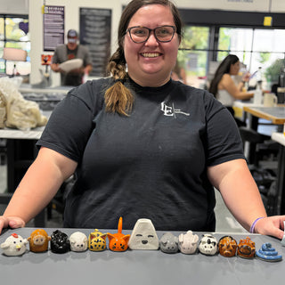 A member standing behind a row of small glazed animal figurines in many colors, ending with a coiled blue snake at The Clay Hole pottery studio in Draper, Utah