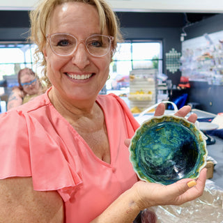 A member holding up a small bowl with a scalloped rim and glossy deep blue and green glaze inside at The Clay Hole pottery studio in Draper, Utah