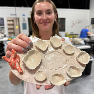 A member holding an oval cream platter ringed with shell shaped dishes, next to a small orange coral piece at The Clay Hole pottery studio in Draper, Utah