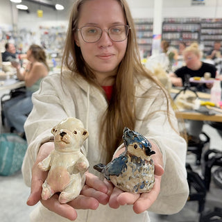 A member holding out two small figures in cupped hands, a cream bear and a blue and gray bird at The Clay Hole pottery studio in Draper, Utah