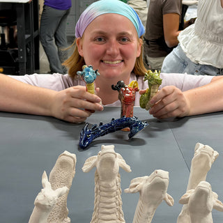 A member holding three small dragon figures in blue, red and green, with white unglazed ones in front at The Clay Hole pottery studio in Draper, Utah