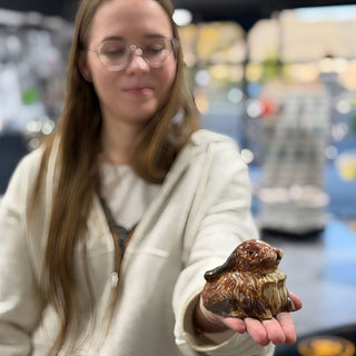 A member holding out a small glossy brown animal figure with a rounded body in an open palm at The Clay Hole pottery studio in Draper, Utah