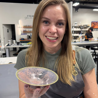 A member holding a small shallow footed dish with a pink and gray mottled glaze and a pale green rim at The Clay Hole pottery studio in Draper, Utah