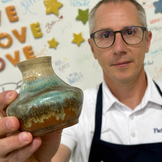 A member holding up a small wide vase with a beige neck and teal and rust drippy glaze at The Clay Hole pottery studio in Draper, Utah