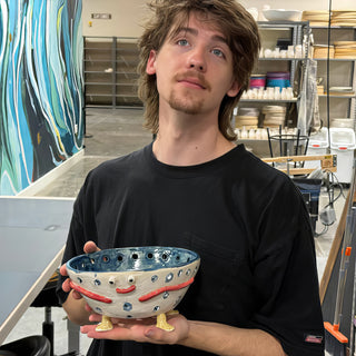 A member holding a small footed bowl with cutout holes, a blue inside, a red smile and tiny yellow feet at The Clay Hole pottery studio in Draper, Utah