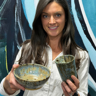A member holding a speckled blue and brown bowl beside a tall rounded vessel with a small hole at The Clay Hole pottery studio in Draper, Utah