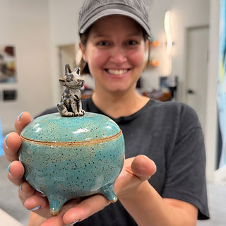 A member holding a round speckled turquoise lidded jar on small feet with a gray animal figure on the lid at The Clay Hole pottery studio in Draper, Utah