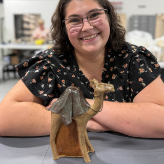 A member smiling with crossed arms behind a tan speckled camel figurine with a gray green blanket on its back at The Clay Hole pottery studio in Draper, Utah