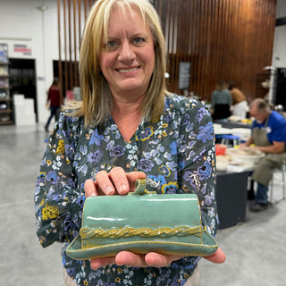 A member holding a teal lidded dish with a loop handle and a braided yellow band along the lid at The Clay Hole pottery studio in Draper, Utah