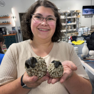 A member holding two small sheep figurines with nubby speckled cream bodies and glossy black heads at The Clay Hole pottery studio in Draper, Utah