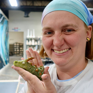 A member holding a tiny green dragon figure with textured scales and orange horns in an open palm at The Clay Hole pottery studio in Draper, Utah