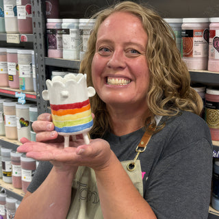 A member holding a tiny white figure with a scalloped top, rainbow stripes, two small ears and short legs at The Clay Hole pottery studio in Draper, Utah