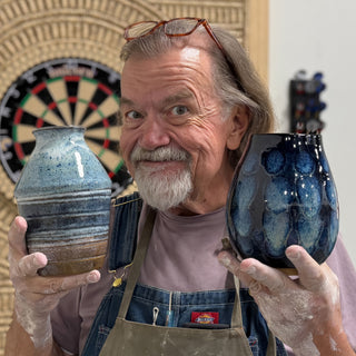 A member holding two vases, one pale blue with horizontal bands and one glossy dark blue with lighter blue spots at The Clay Hole pottery studio in Draper, Utah