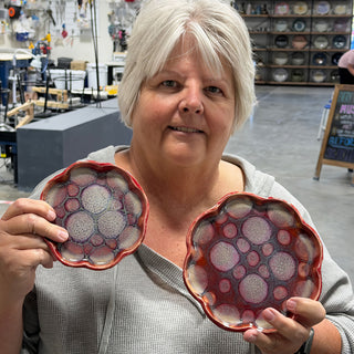A member holding two small scalloped dishes with red rims and a pattern of pale and pink bubbles at The Clay Hole pottery studio in Draper, Utah