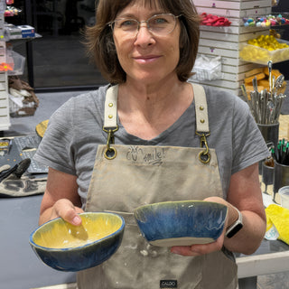 A member holding two small bowls, one yellow inside and dark blue outside, the other streaked blue, with The Clay Hole, a pottery studio in Draper, Utah