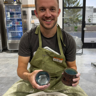 A member holding two small cups, a blue gray one and a dark brown one, both with teal glaze inside at The Clay Hole pottery studio in Draper, Utah