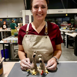 A member in a tan apron with two small clay creatures with long necks and open mouths among tiny starfish at The Clay Hole pottery studio in Draper, Utah