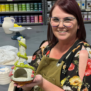 A member holding a tall cone shaped sculpture with an arched opening, green vines and small blue flowers at The Clay Hole pottery studio in Draper, Utah
