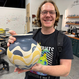 A member holding a round vase with wavy blue-gray and yellow bands on a cream body at The Clay Hole pottery studio in Draper, Utah