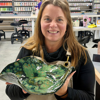 A member holding a wavy edged platter in glossy dark green glaze with light green spots and swirls at The Clay Hole pottery studio in Draper, Utah