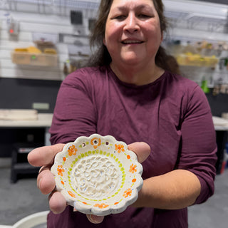 A member holding a small white scalloped dish with a textured center, orange flowers and a green dotted ring at The Clay Hole pottery studio in Draper, Utah