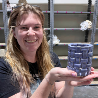A member holding up a small cup with open basket weave sides and a glossy periwinkle purple glaze at The Clay Hole pottery studio in Draper, Utah