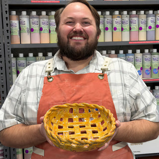 A member in an orange apron holding a woven yellow-orange bowl with a braided rim and open lattice sides at The Clay Hole pottery studio in Draper, Utah