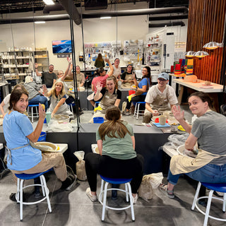 A class of potters sits on blue stools at pottery wheels, turning to wave at the camera at The Clay Hole in Draper, Utah