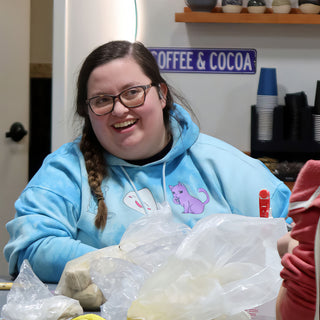 A smiling potter in a blue tie-dye hoodie sits behind plastic bags of clay at a table at The Clay Hole in Draper, Utah