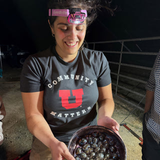 A smiling person in a pink headlamp holds out a metal bowl filled with small shiny dark blue spheres at The Clay Hole in Draper, Utah