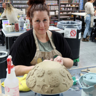 A smiling potter rests hands on an upside-down tan clay bowl covered with raised flower and starfish shapes at The Clay Hole in Draper, Utah