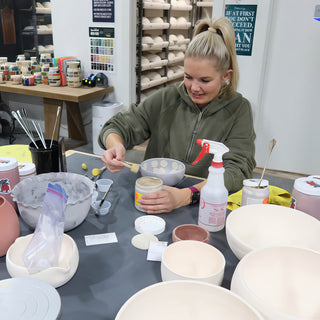 A potter in an olive hoodie paints a small gray bowl with a brush, surrounded by white bowls and jars at The Clay Hole in Draper, Utah