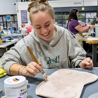 A smiling potter brushing pale pink color onto a flat clay slab at a worktable beside a jar at The Clay Hole in Draper, Utah