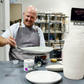 A smiling potter brushes a pale green plate beside a tall white vase with a ridged texture at The Clay Hole in Draper, Utah