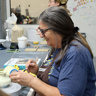 A potter smiling while brushing detail onto a small pale green vase with a blue band near the rim at The Clay Hole in Draper, Utah