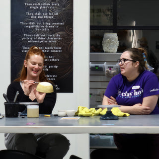 A potter smiling while brushing a pale yellow bowl at a table, with a second potter in purple nearby at The Clay Hole in Draper, Utah