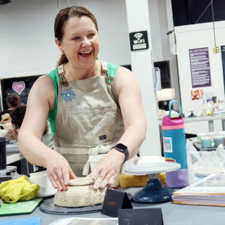 A smiling potter pressing both hands onto a rounded clay piece with carved swirl patterns on a turntable at The Clay Hole in Draper, Utah