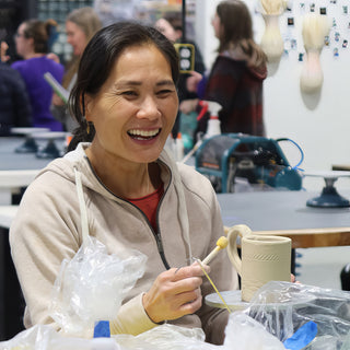 A potter smiling with a thin yellow-tipped tool beside an unglazed handled cup with carved bands on a turntable at The Clay Hole in Draper, Utah