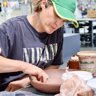 A potter in a green cap etches a pattern into the side of a wide reddish brown bowl at The Clay Hole in Draper, Utah