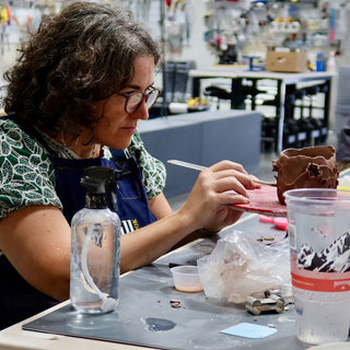A potter in glasses works a thin tool over a small textured brown clay piece, with a spray bottle nearby at The Clay Hole in Draper, Utah