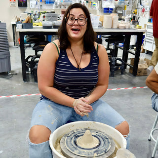 A potter with an open-mouthed grin sits at a pottery wheel topped with a small pale clay cone at The Clay Hole in Draper, Utah