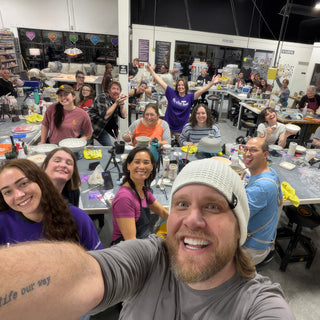 A class of potters at long worktables smiling and cheering, with a person in a white beanie taking a selfie at The Clay Hole in Draper, Utah