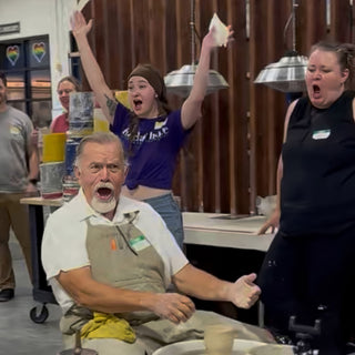 A seated potter at a pottery wheel and others around them cheer with open mouths and one arm raised high at The Clay Hole in Draper, Utah