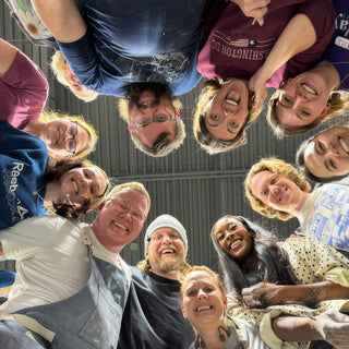 A ring of smiling potters leaning over the camera, a few in aprons and one in a gray beanie at The Clay Hole in Draper, Utah